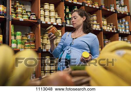 Stock Photo - A pregnant woman considering a jar of vegetables at the supermarket. Fotosearch