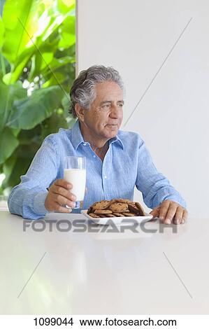 A senior man sitting at a table with chocolate chip cookies and a glass of milk View Large Photo Image Picture - A senior man sitting at a table with chocolate chip cookies and a glass of milk. Fotosearch