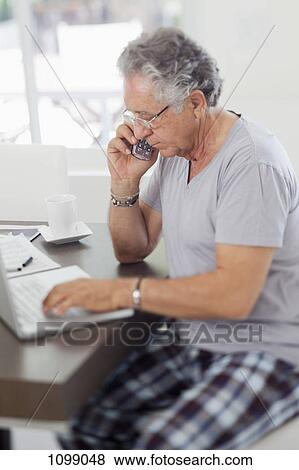 A senior man using a laptop and talking on a cordless phone at home View Large Photo Image Stock Photo - A senior man using a laptop and talking on a cordless phone at home. Fotosearch