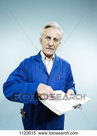 Stock Photography - A senior man wearing coveralls and pointing to a paperwork on a clipboard. Fotosearch