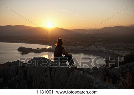 Stock Image - A silhouetted person sitting atop a hill watching the sunset, Rhodes, Greece. Fotosearch