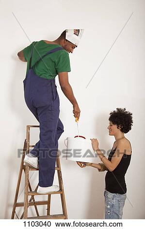 Stock Image - A woman holding a bucket for a man painting a wall with a paint roller. Fotosearch