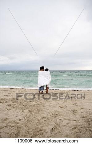 A young couple wrapped in a beach towel standing on the beach View Large Photo Image Stock Photograph - A young couple wrapped in a beach towel standing on the beach. Fotosearch