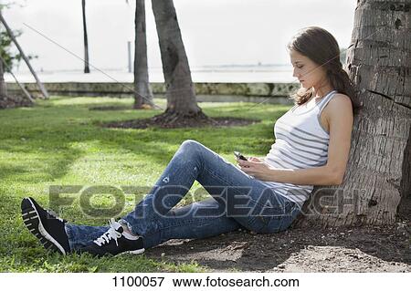 Stock Photo - Girl relaxing against tree and using mobile phone. Fotosearch