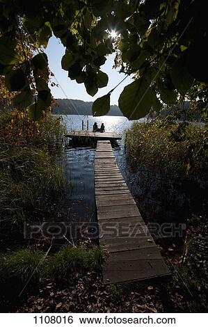 Stock Photograph - Two people sitting at the end of a jetty, silhouetted. Fotosearch