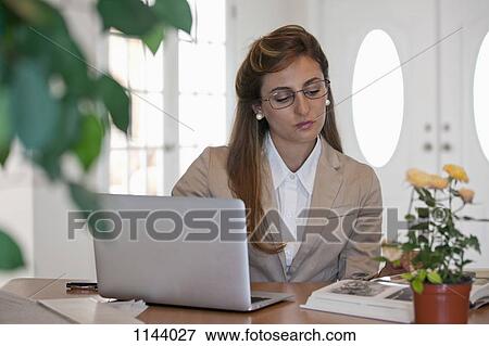 Stock Photo - A businesswoman using a laptop and reading a book at her office desk. Fotosearch