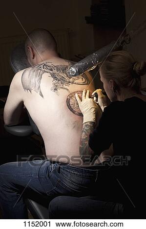 A female tattoo artist tattooing a man's back in a tattoo shop View Large Photo Image Stock Image - A female tattoo artist tattooing a man's back in a tattoo shop. Fotosearch