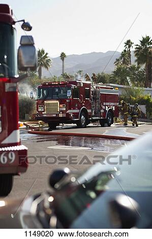 Stock Image - A fire truck and firefighters on a suburban street next to a burning house. Fotosearch