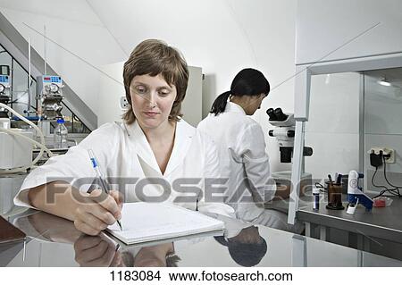 A lab technician writing notes while another technician works in background View Large Photo Image Picture - A lab technician writing notes while another technician works in background. Fotosearch