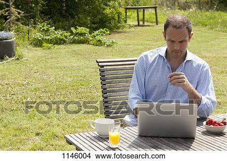 A man sitting at a table in his backyard having breakfast and using a laptop View Large Photo Image Picture - A man sitting at a table in his backyard having breakfast and using a laptop. Fotosearch