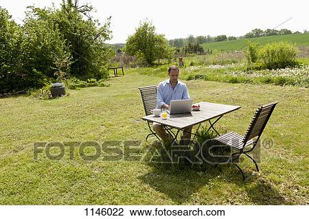 A man sitting at a table in his backyard having breakfast and using a laptop View Large Photo Image Stock Image - A man sitting at a table in his backyard having breakfast and using a laptop. Fotosearch