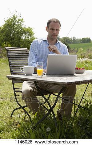 A man sitting at a table in his backyard having breakfast and using a laptop View Large Photo Image Stock Photograph - A man sitting at a table in his backyard having breakfast and using a laptop. Fotosearch
