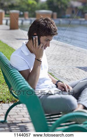 A man sitting on a park bench talking on a mobile and using a laptop View Large Photo Image Stock Photo - A man sitting on a park bench talking on a mobile and using a laptop. Fotosearch