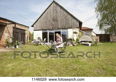 A woman sitting at a table in her backyard using a laptop View Large Photo Image Stock Photo - A woman sitting at a table in her backyard using a laptop. Fotosearch