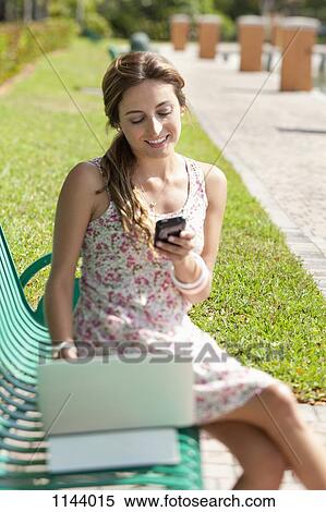 A woman sitting on a park bench using a laptop and a mobile phone View Large Photo Image Stock Photography - A woman sitting on a park bench using a laptop and a mobile phone. Fotosearch