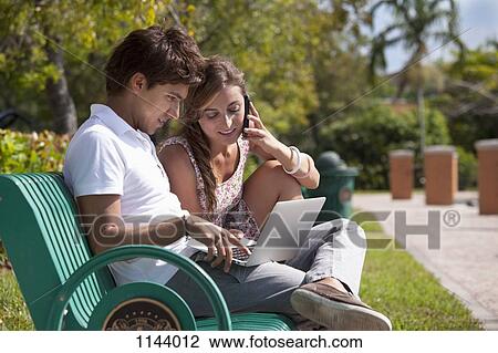A young couple sitting on a park bench using a laptop and mobile phone View Large Photo Image Stock Image - A young couple sitting on a park bench using a laptop and mobile phone. Fotosearch