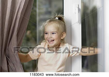 Stock Image - A young laughing girl pushing window curtains apart. Fotosearch