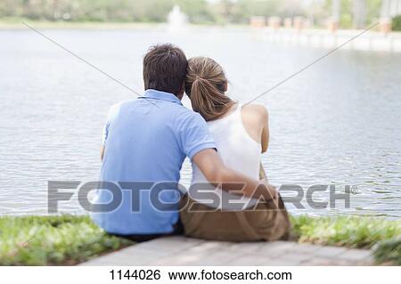 Stock Photograph - An affectionate couple sitting by a lake, rear view. Fotosearch