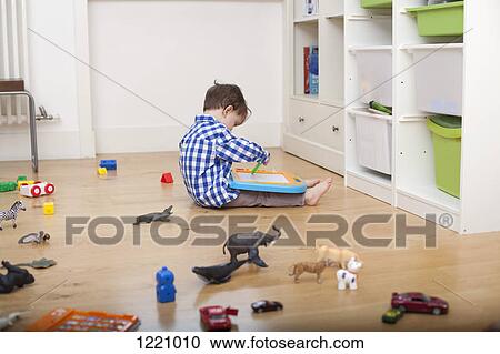 Stock Image - A boy sitting on the floor, drawing on a tablet, surrounded by various toys. Fotosearch