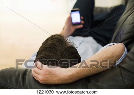 Stock Image - A man lying on a sofa looking at smart phone, view from top of head. Fotosearch