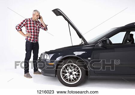 Stock Image - A man standing next to a broke down car while using a cell phone. Fotosearch