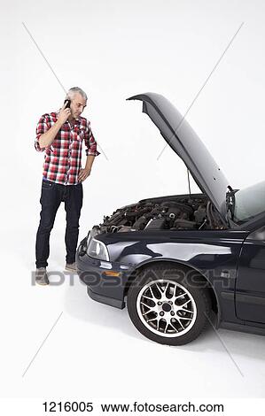 Stock Photography - A man standing next to a broke down car while using a cell phone. Fotosearch