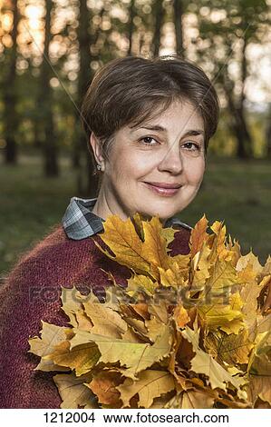 Picture - A mature woman holding a bunch of autumn leaves in nature. Fotosearch
