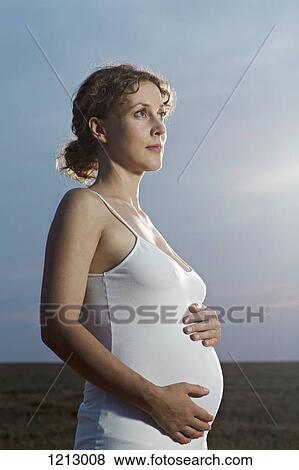 Stock Photo - A pregnant woman holding her belly and looking away thoughtfully. Fotosearch