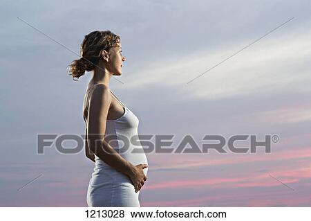 Stock Photo - A pregnant woman holding her belly and looking away at a dramatic sky. Fotosearch