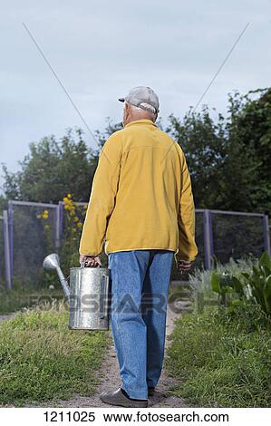 Stock Photography - A senior man carrying a metal water can in his backyard. Fotosearch