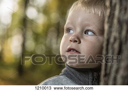 Stock Image - A young boy leaning against a tree trunk looking away thoughtfully. Fotosearch