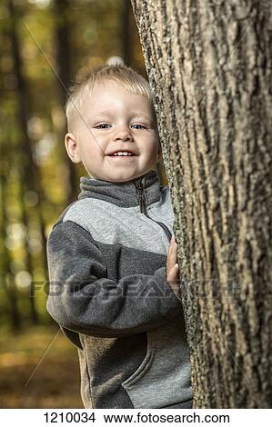 Picture - A young boy standing by a tee trunk and smiling happily. Fotosearch