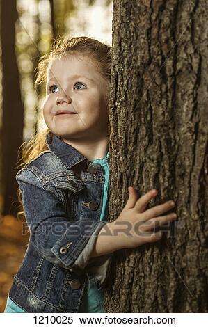 A young girl holding on to a tree trunk while looking away curiously View Large Photo Image Stock Photography - A young girl holding on to a tree trunk while looking away curiously. Fotosearch