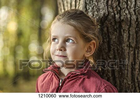 A young girl looking away curiously while leaning against a tree trunk View Large Photo Image Stock Image - A young girl looking away curiously while leaning against a tree trunk. Fotosearch