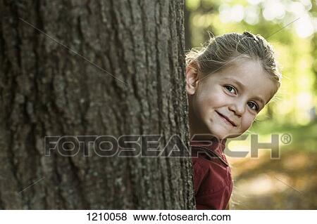 A young smiling girl peeking from behind a tree trunk View Large Photo Image Stock Photo - A young smiling girl peeking from behind a tree trunk. Fotosearch