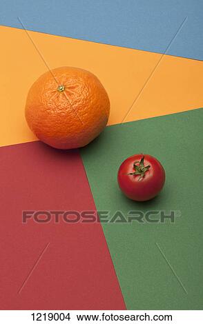 Picture - An orange and a tomato placed on a multi colored geometric pattern. Fotosearch