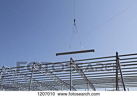 Stock Image - Crane lowering steel beam towards construction frame. Fotosearch