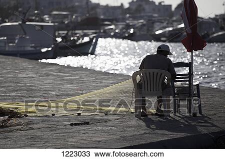Stock Image - Fisherman on seat by harbor preparing nets. Fotosearch