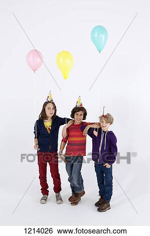 Stock Photograph - Three boys wearing party hats and holding balloons, in a row. Fotosearch
