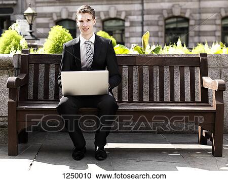 A cheerful young businessman using a laptop while sitting outdoors on a bench View Large Photo Image Stock Image - A cheerful young businessman using a laptop while sitting outdoors on a bench. Fotosearch