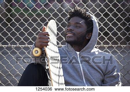 A grinning young man leaning against a chain link fence holding a skateboard View Large Photo Image Stock Photograph - A grinning young man leaning against a chain link fence holding a skateboard. Fotosearch