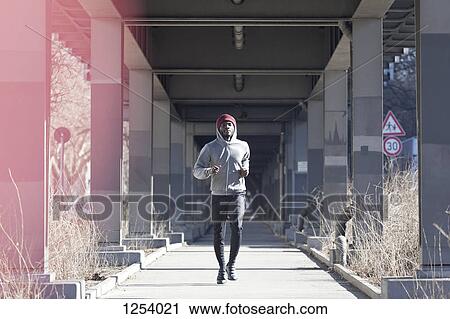 Stock Image - A hip young man jogging on a city sidewalk under a bridge. Fotosearch