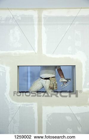 Stock Image - A house painter holding a trowel, seen through window hole in a newly constructed wall. Fotosearch