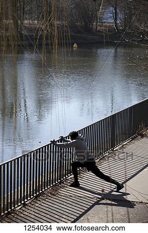 A man doing a stretching lunge against a railing near a river View Large Photo Image Picture - A man doing a stretching lunge against a railing near a river. Fotosearch