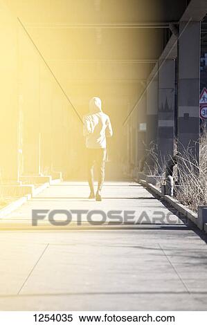 Stock Photography - A man jogging on a city sidewalk under a bridge, rear view. Fotosearch