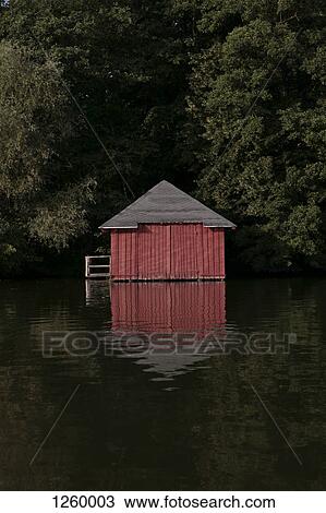 Stock Image - A red boathouse nestled amongst trees on a lake. Fotosearch