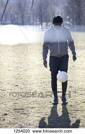 Stock Image - A young man kicking a soccer ball in a city park. Fotosearch