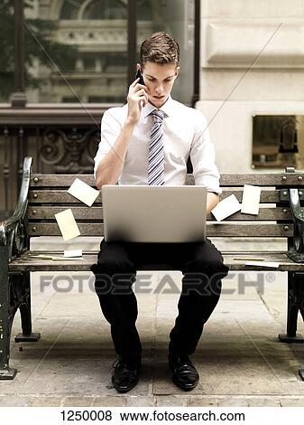 Stock Photo - Businessman using a smart phone while using a laptop and sitting on bench with sticky notes. Fotosearch
