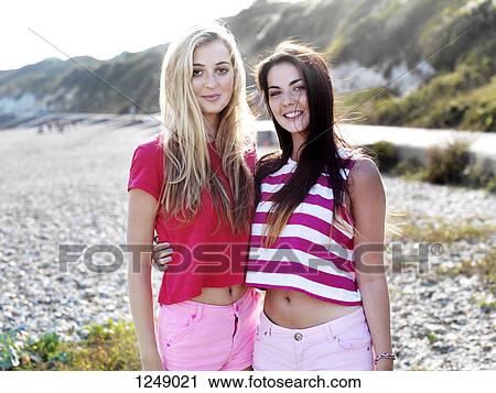 Stock Image - Two smiling young women with their arms around each other standing on a beach. Fotosearch