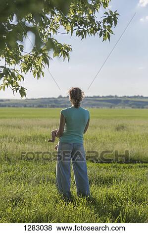 Stock Photo - A mother standing in a field while holding her baby and looking at the view . Fotosearch